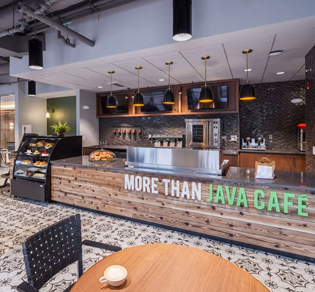 Interior view of a modern café counter with a wooden front displaying the text 'MORE THAN JAVA CAFÉ'. The counter has a display case with pastries, a coffee machine, and several hanging pendant lights above. A round wooden table with a cup of coffee and a black woven chair are visible in the foreground. The floor has a patterned tile design.