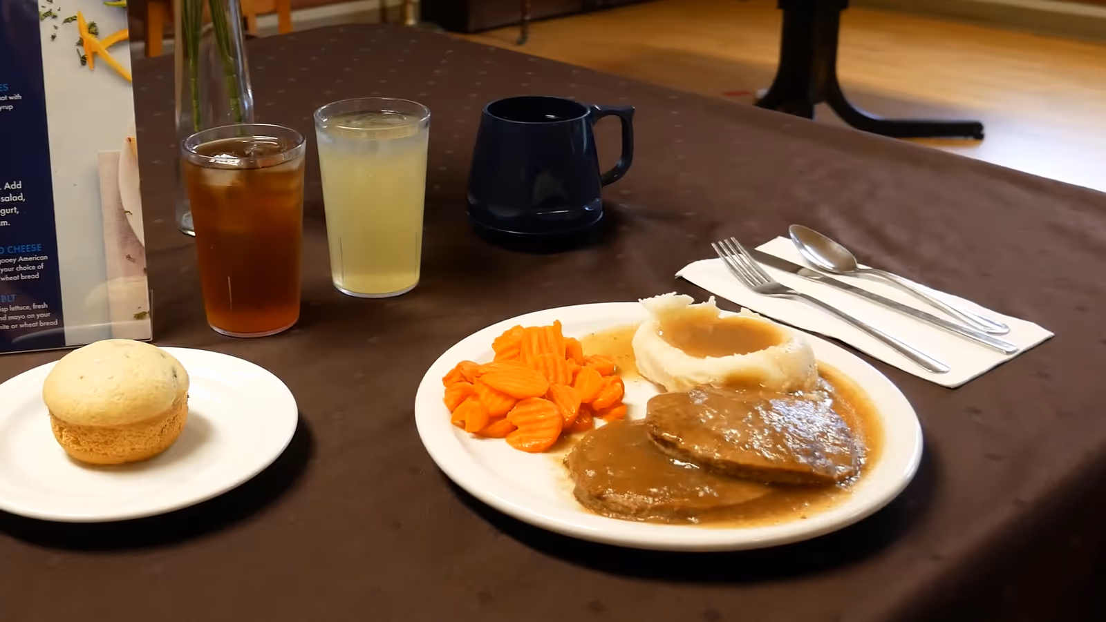 A plated meal of sliced roast with gravy, mashed potatoes, carrots, a roll, and drinks on a brown tablecloth in a dining area.