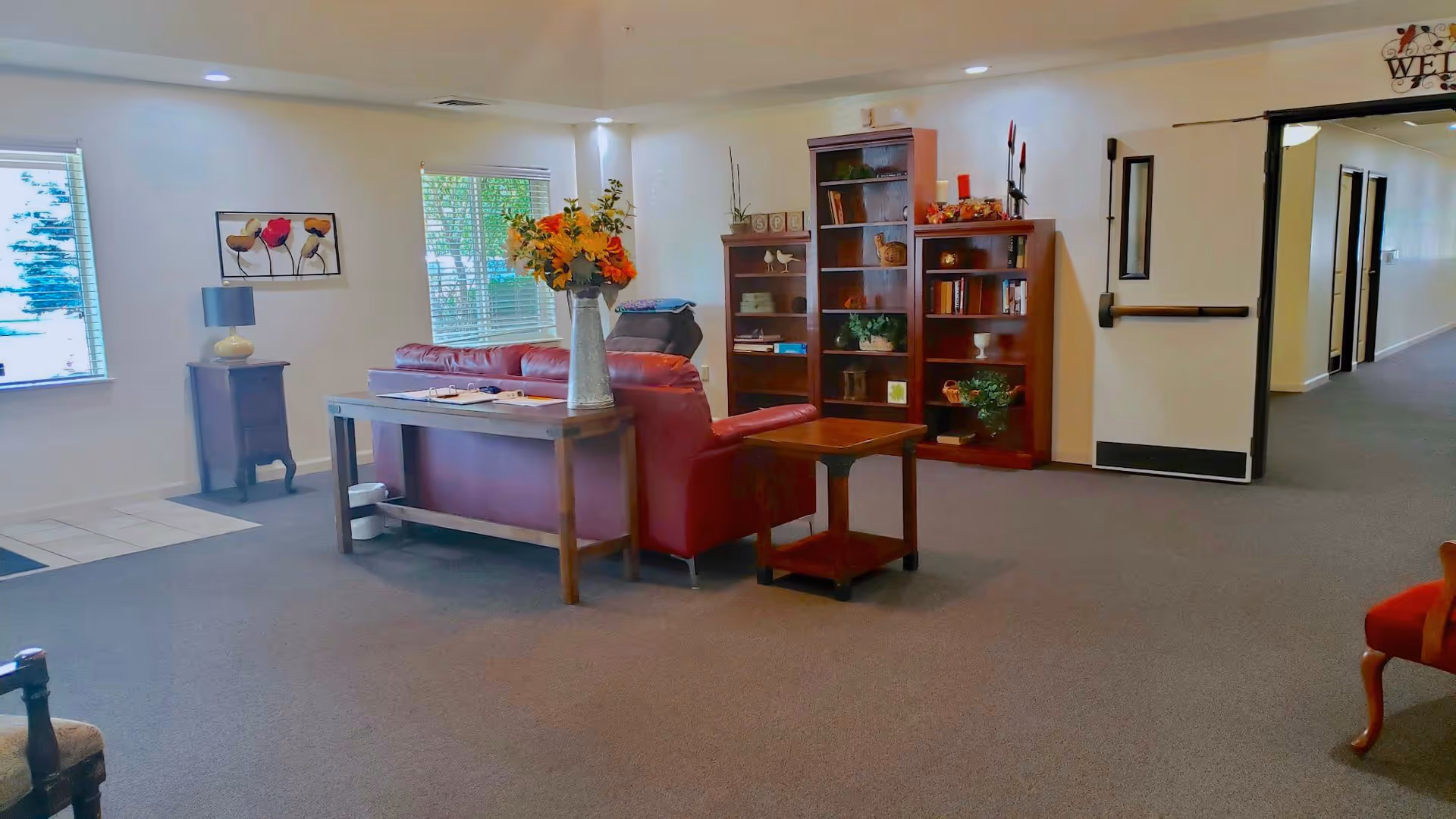 A cozy living room area in a senior living facility with a red leather sofa, wooden side tables, a vase with flowers, bookshelves filled with books and decorative items, and a hallway with open double doors leading to other rooms.