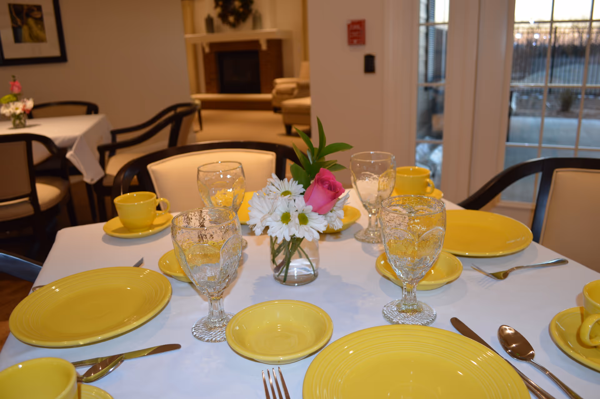 A dining table set with yellow plates, cups, and saucers, along with clear water glasses and silverware. A small vase with white daisies and a pink rose is placed in the center of the table. In the background, there are more tables and chairs, a fireplace, and large windows letting in natural light.