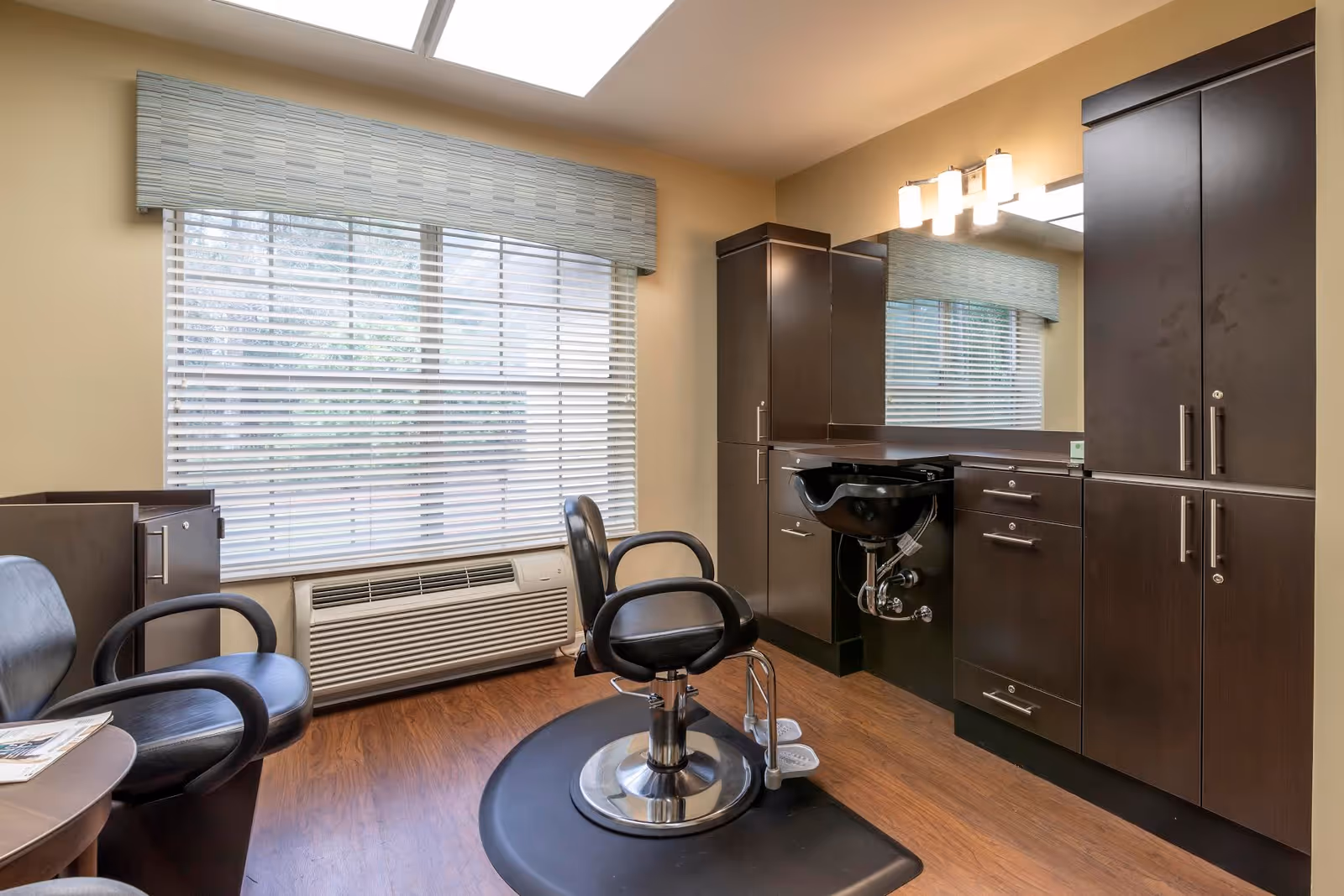 Interior salon room with a styling chair, shampoo sink, dark cabinets and a large window with blinds.