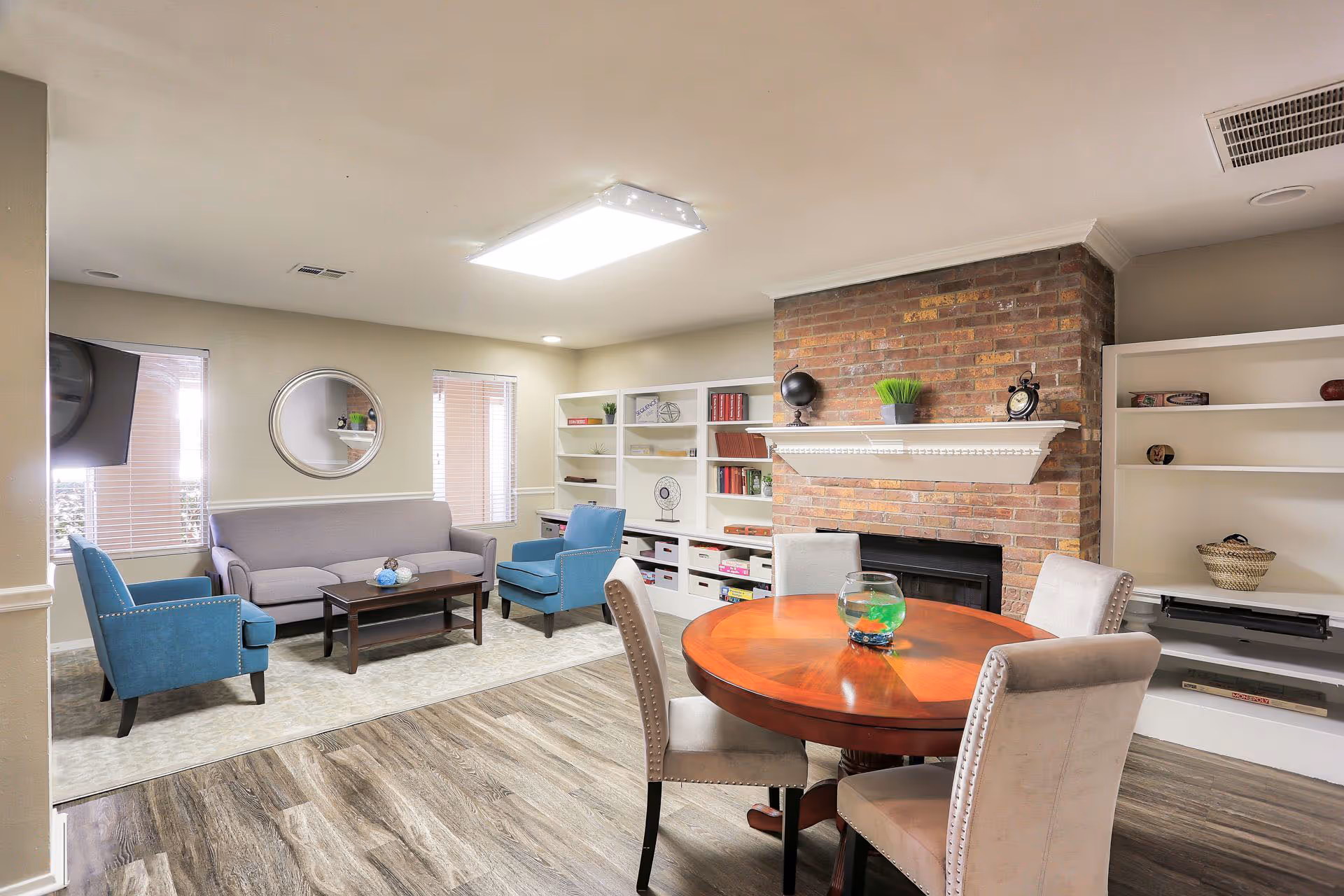 A bright communal living room with sofas and armchairs, a round wooden table, brick fireplace and built-in shelving.
