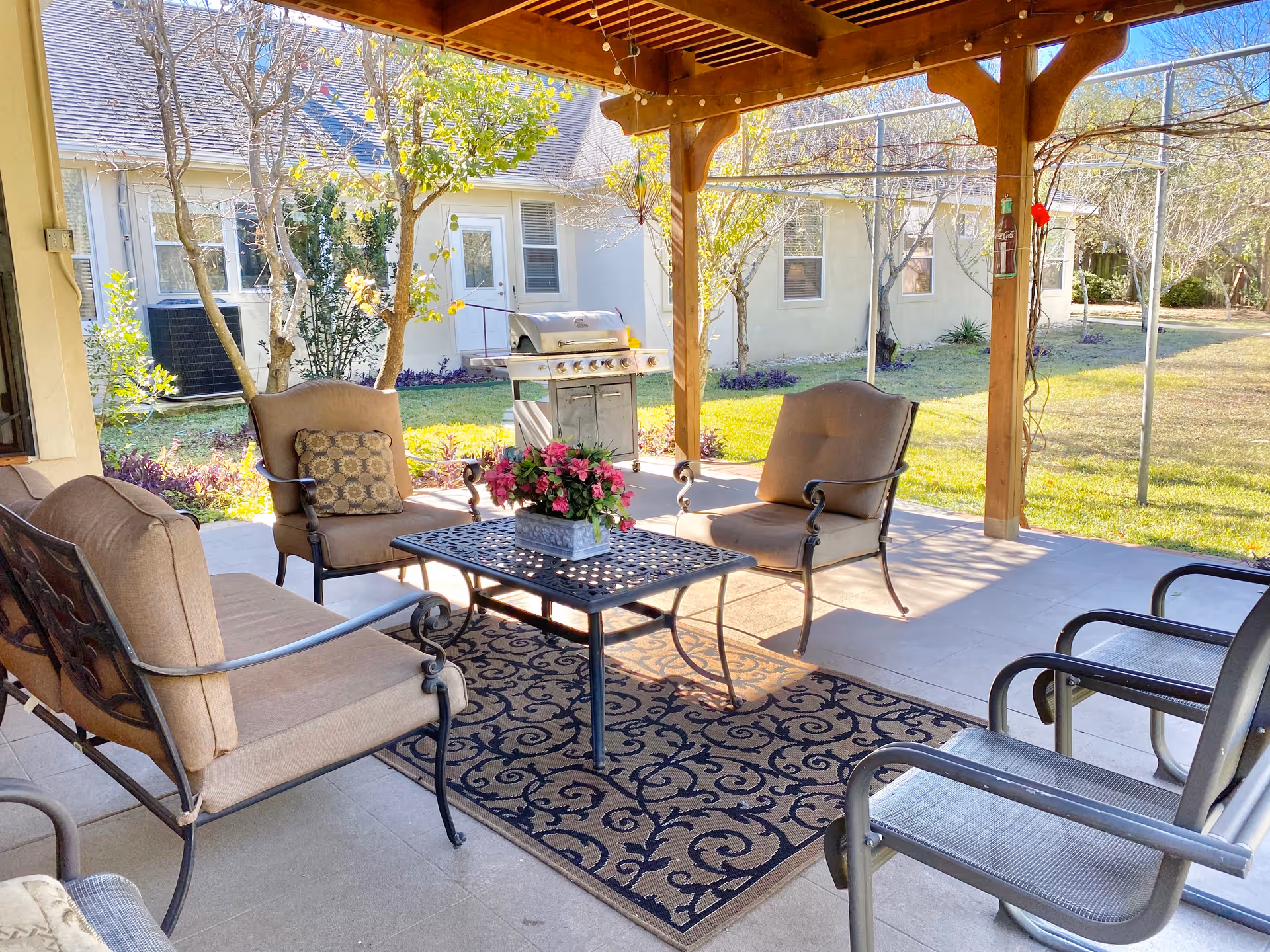 Outdoor covered patio area with cushioned chairs and a loveseat arranged around a metal coffee table with a flower pot centerpiece. A patterned rug is underneath the table. In the background, there is a barbecue grill, trees, and a building with windows and a door.