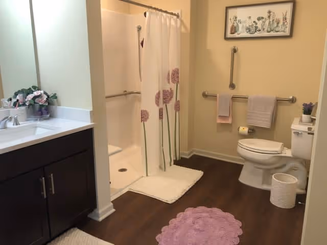 A bathroom with a walk-in shower featuring a white curtain with purple flower designs, a white toilet with grab bars on the wall, a dark wood vanity with a white countertop and sink, and several bath mats in white and purple. There is a framed floral artwork above the toilet and a small plant on the toilet tank.