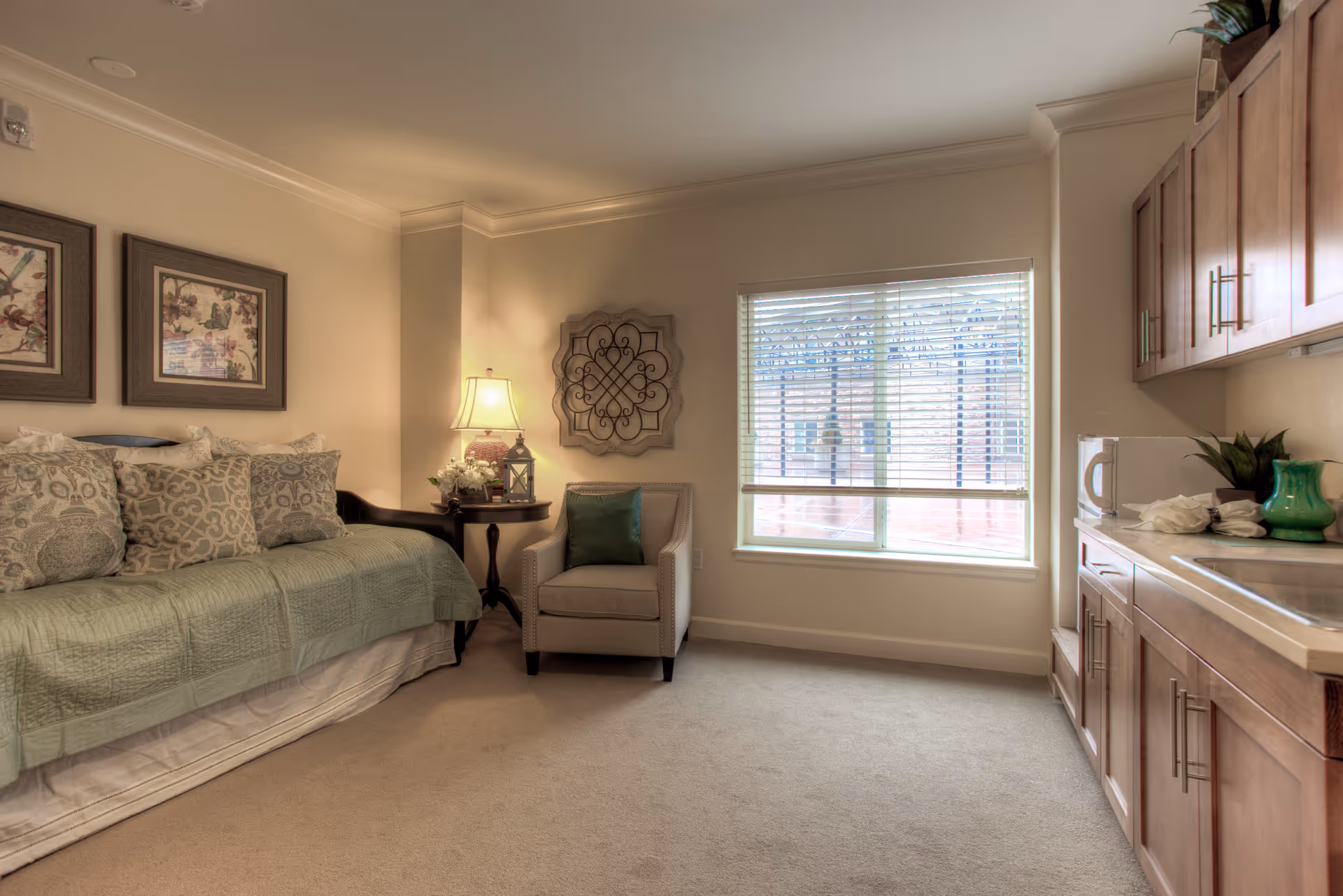 A cozy room in a retirement community featuring a daybed with multiple patterned pillows, a side table with a lamp and decorative items, an armchair with a green cushion, a window with blinds letting in natural light, and a kitchenette area with wooden cabinets, a microwave, and a green vase.