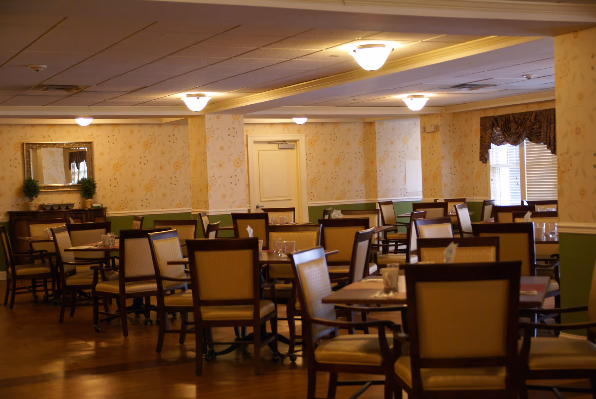 A well-lit dining room with multiple wooden tables and cushioned chairs arranged throughout the room.