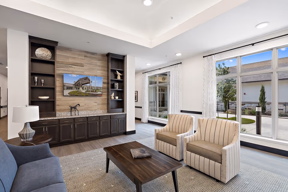 A bright and modern living room area in a senior living facility featuring a blue sofa, two beige striped armchairs, a dark wooden coffee table with a book on it, and a built-in dark wood cabinet with shelves displaying decorative items. Large windows with white curtains allow natural light to fill the room, and a flat-screen TV mounted on a wood-paneled wall shows an exterior view of the facility.