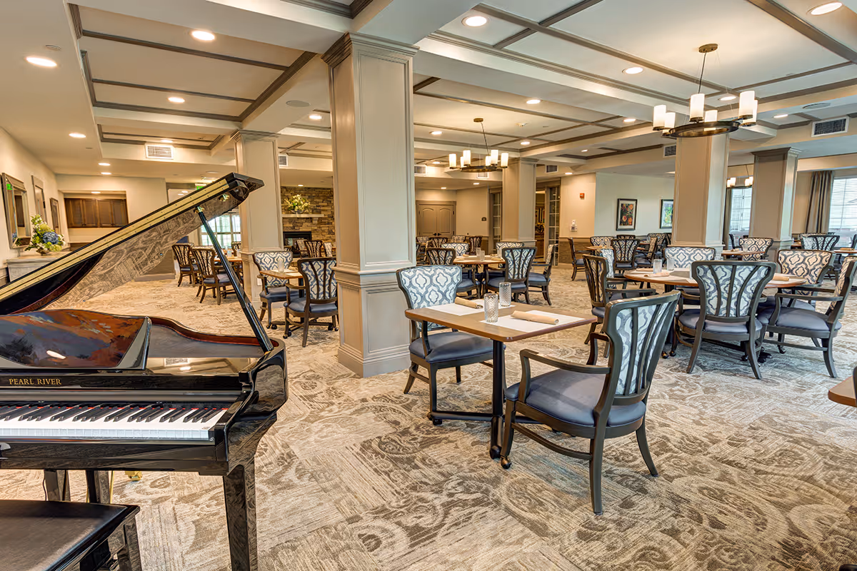 Spacious, well-lit dining room with multiple tables and patterned chairs, a grand piano in the foreground, and columns and chandeliers throughout.