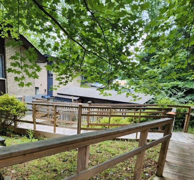 A wooden ramp with railings surrounded by greenery and trees, leading to a building with light-colored brick walls and dark trim, under a canopy of green leaves.