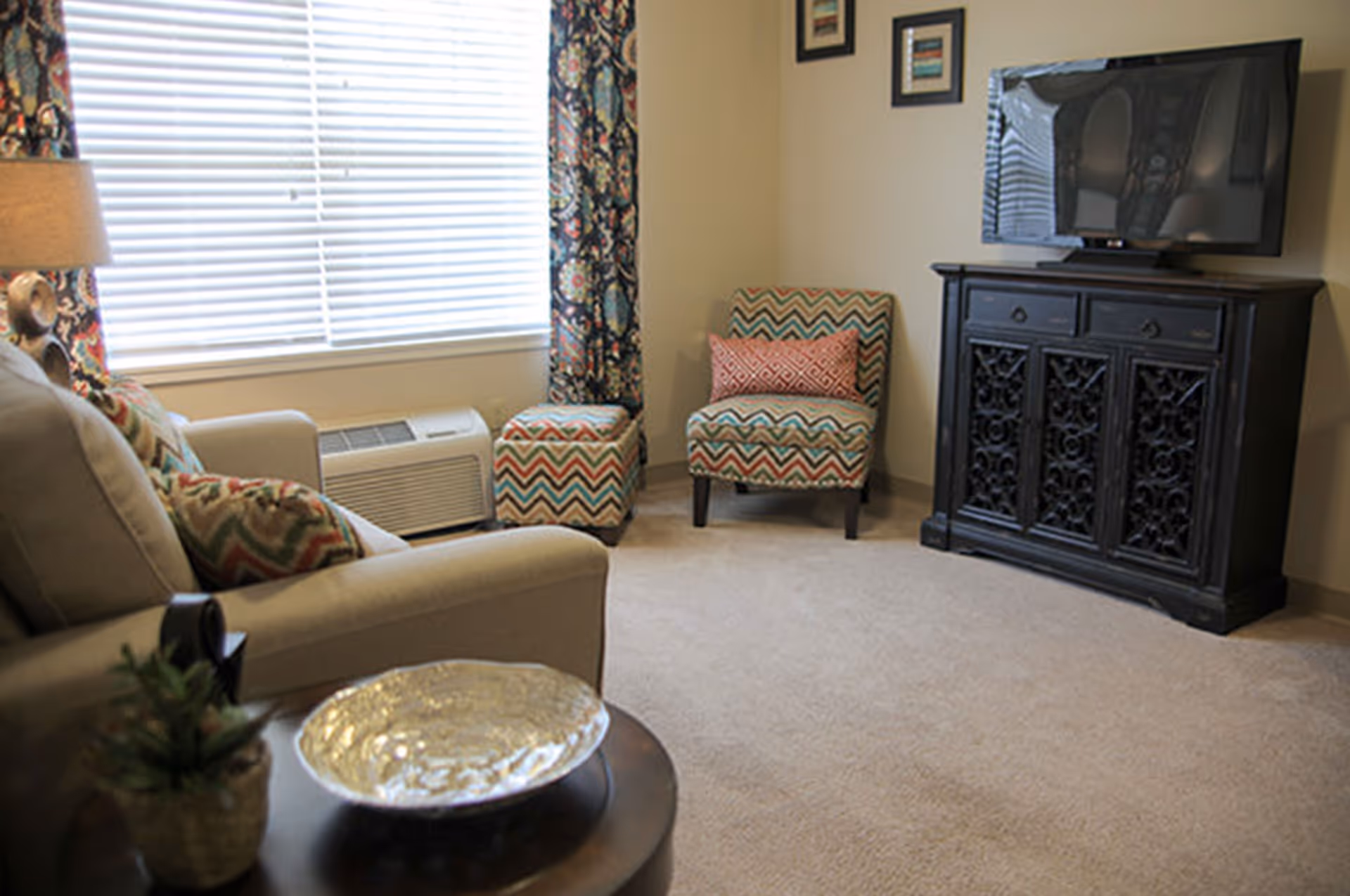 Cozy living room with a sofa, patterned chairs and ottoman, a TV on a decorative cabinet, and a window with blinds.