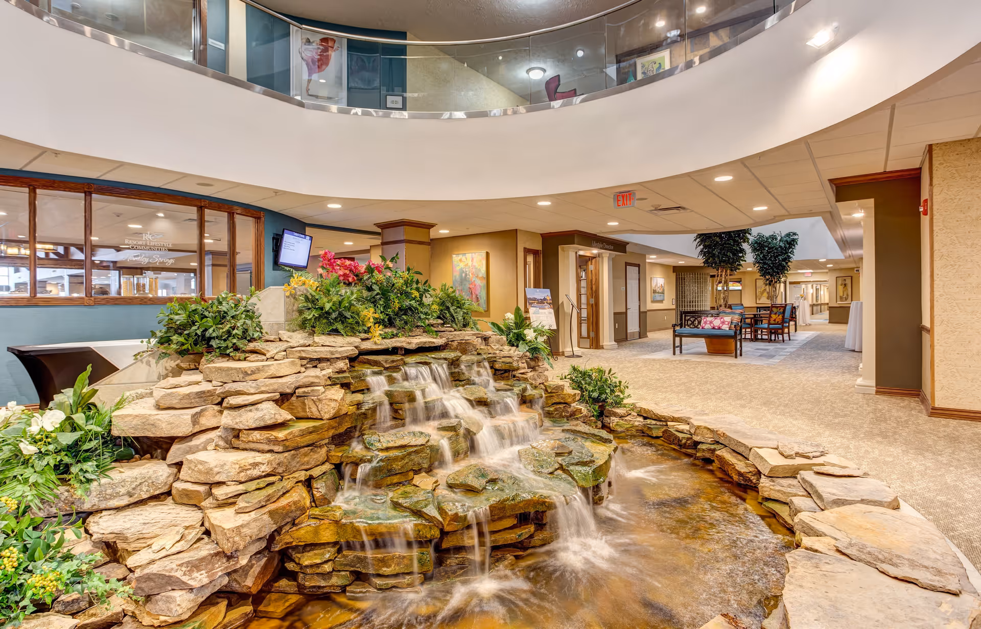 Indoor area of a senior living facility featuring a decorative rock waterfall with flowing water surrounded by plants. The space has carpeted floors, seating areas with chairs and tables, and warm lighting. There is a curved balcony with glass railing above and various artworks on the walls.