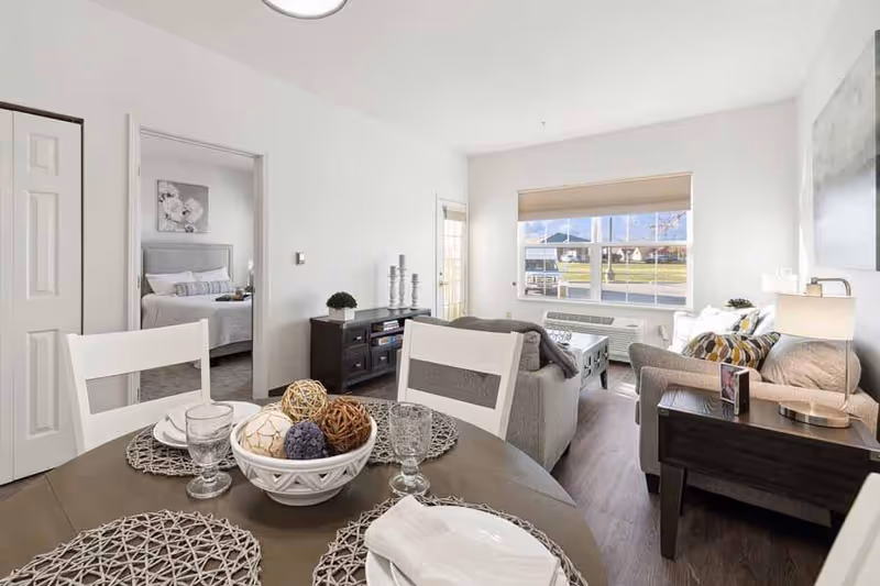 A bright and modern living and dining area in a senior living facility. The dining table is set with placemats, plates, glasses, and a decorative bowl with woven balls. The living room has a sofa, armchair, side table with a lamp, and a TV stand. A large window lets in natural light, and a bedroom is visible through an open door.