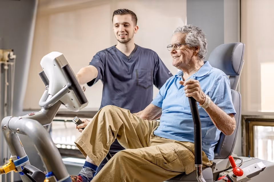 An elderly man wearing glasses and a light blue polo shirt is exercising on a recumbent stationary bike while a young male healthcare worker in navy scrubs assists and encourages him in a rehabilitation center.