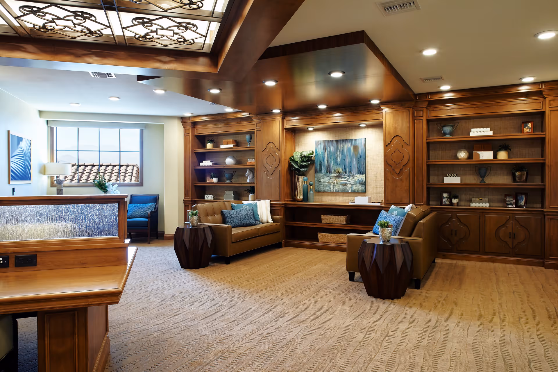 A spacious and well-lit living room area with two brown leather sofas adorned with blue and white cushions. The room features built-in wooden bookshelves filled with decorative items and books, a large window letting in natural light, and a wooden ceiling with recessed lighting. There are two small wooden side tables with potted plants, and a large abstract painting on the back wall.