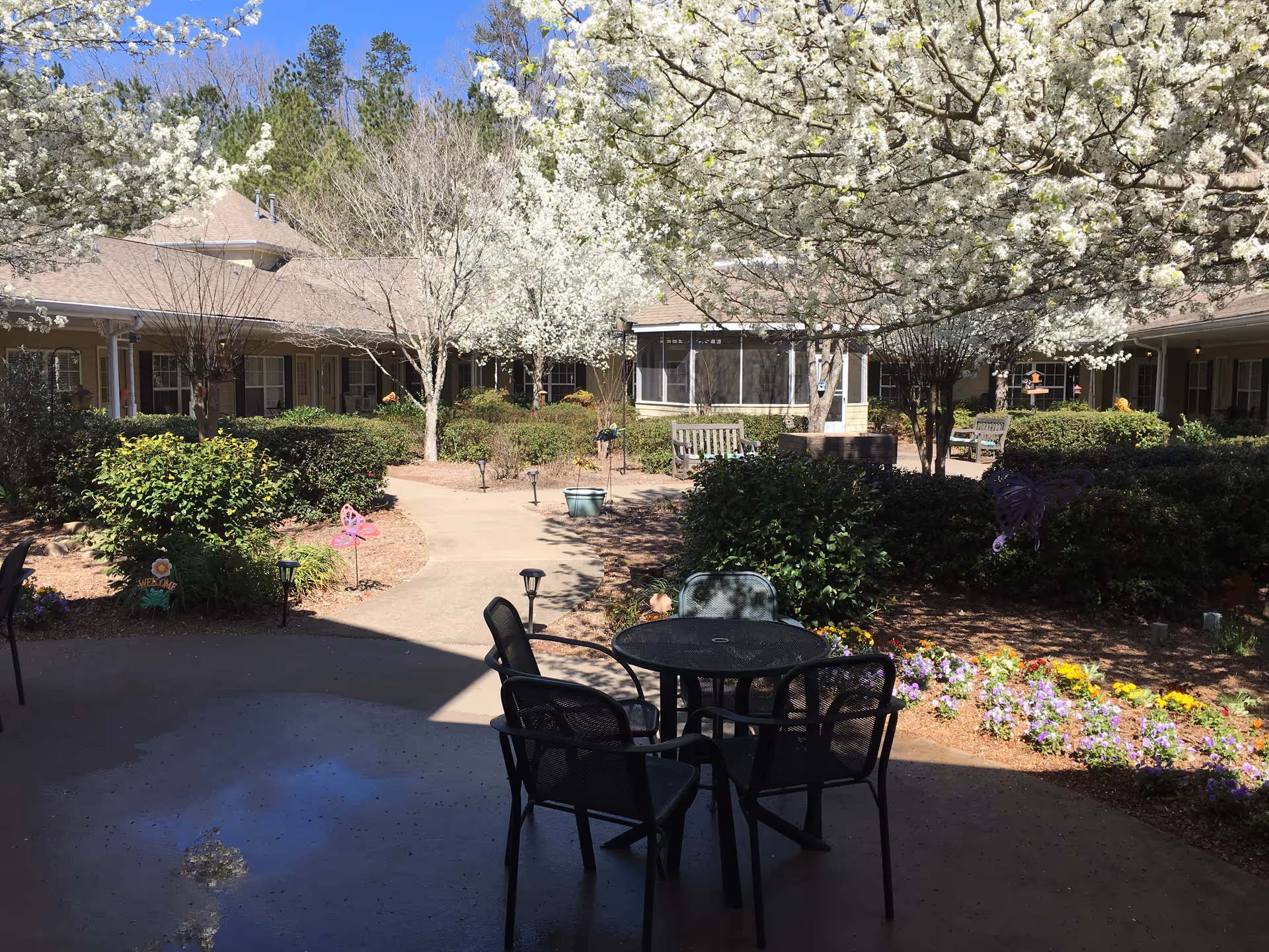 Outdoor courtyard area at a senior living facility with a round black metal table and four chairs in the foreground. The courtyard features blooming white flowering trees, colorful flower beds, green bushes, and a paved walkway leading to a building with a beige roof and multiple windows. Wooden benches are placed along the walkway under the trees.