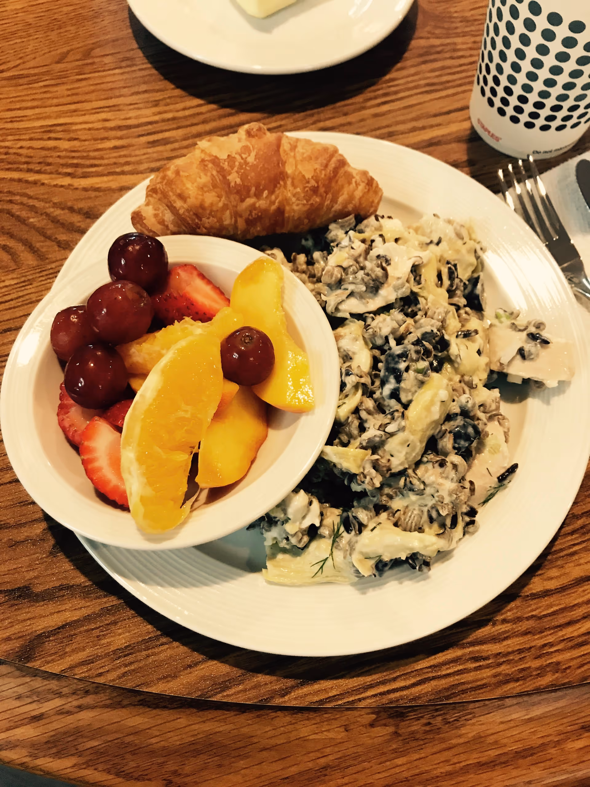 A white plate on a wooden table with a croissant, a serving of creamy wild rice salad, and a small bowl of mixed fruit including grapes, strawberries, orange slices, and peach slices. A cup with a black dot pattern and a fork and knife are partially visible beside the plate.