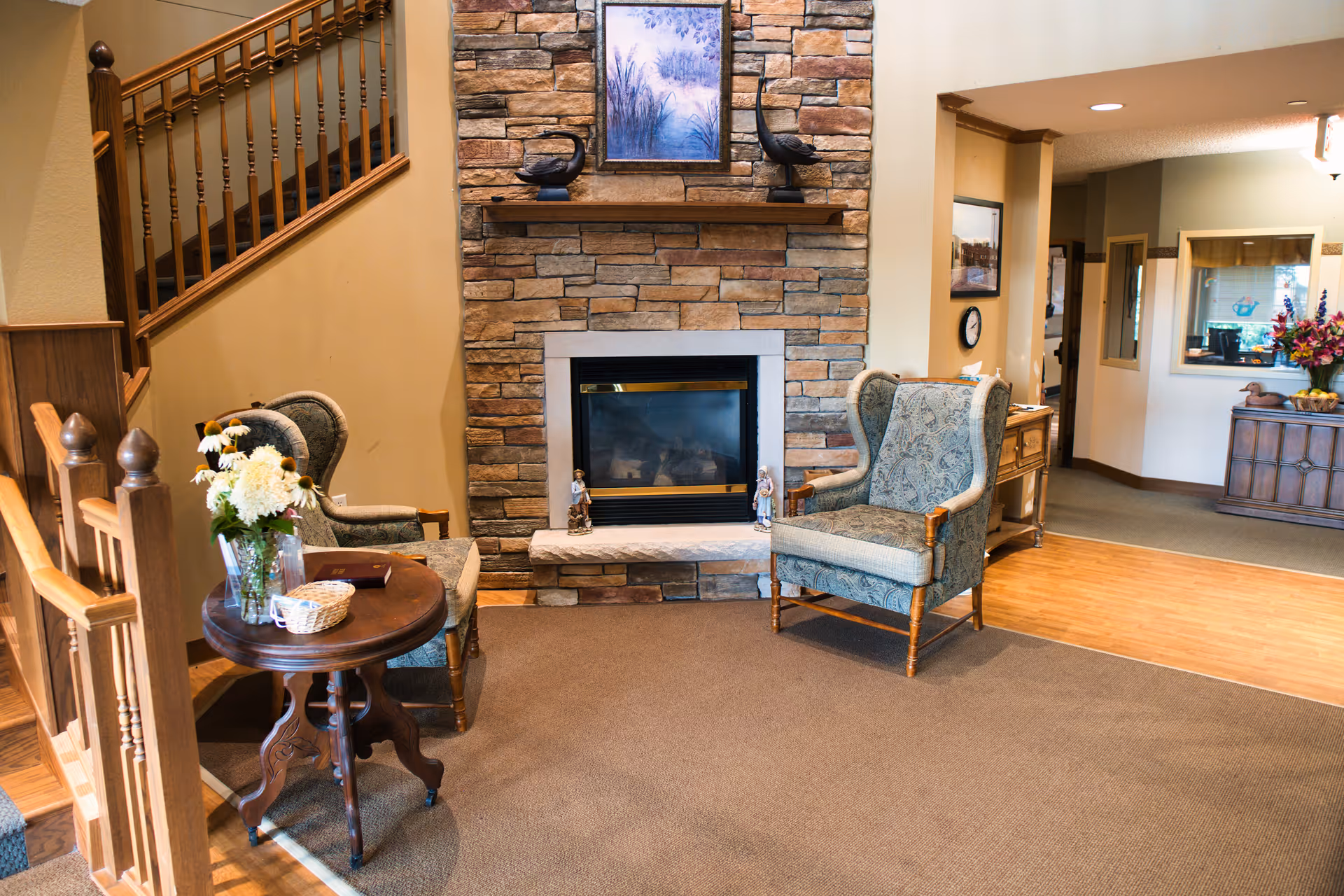 A cozy sitting area in an assisted living facility featuring a stone fireplace with a framed picture above it, two upholstered armchairs, a small round wooden table with a vase of flowers and a basket, wooden staircase railing on the left, and a hallway leading to other rooms on the right.