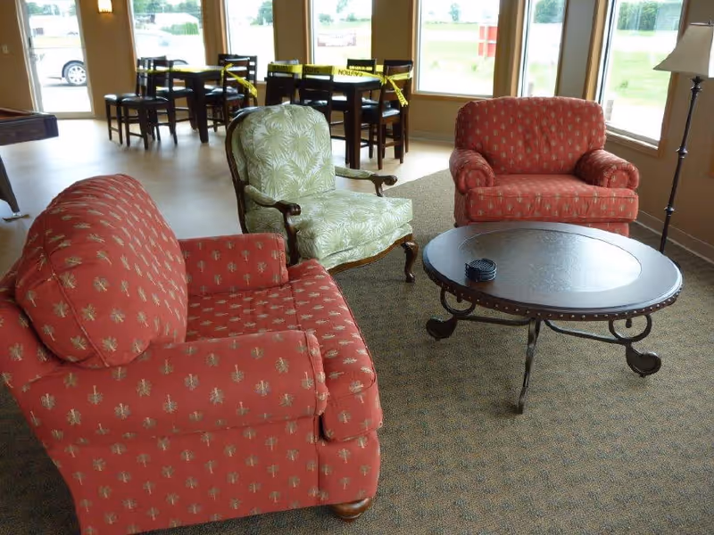 A seating area with two red patterned armchairs and one green patterned armchair arranged around a round black coffee table on a carpeted floor. Behind the seating area, there are large windows letting in natural light and a dining area with tables and chairs, some of which are blocked off with yellow caution tape.
