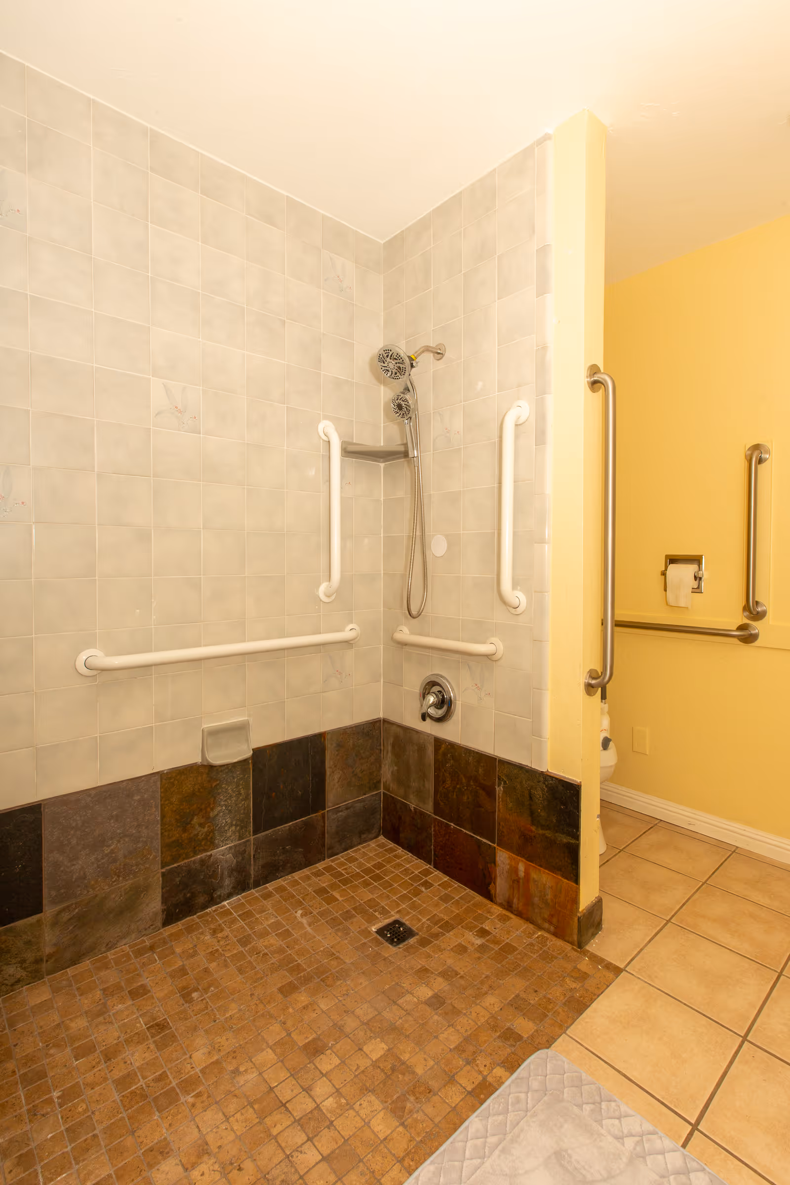 Accessible shower area with beige and brown tiled floor and walls, equipped with multiple white grab bars and a handheld showerhead. Adjacent to the shower is a yellow wall with additional grab bars and a toilet paper holder, indicating a bathroom setup.