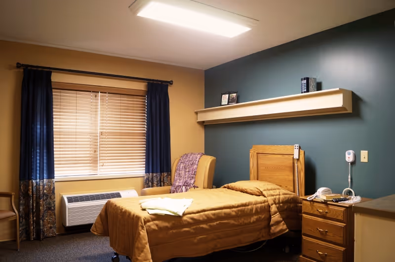 A tidy senior care bedroom with a single bed, nightstand, armchair, and a window with blue curtains.