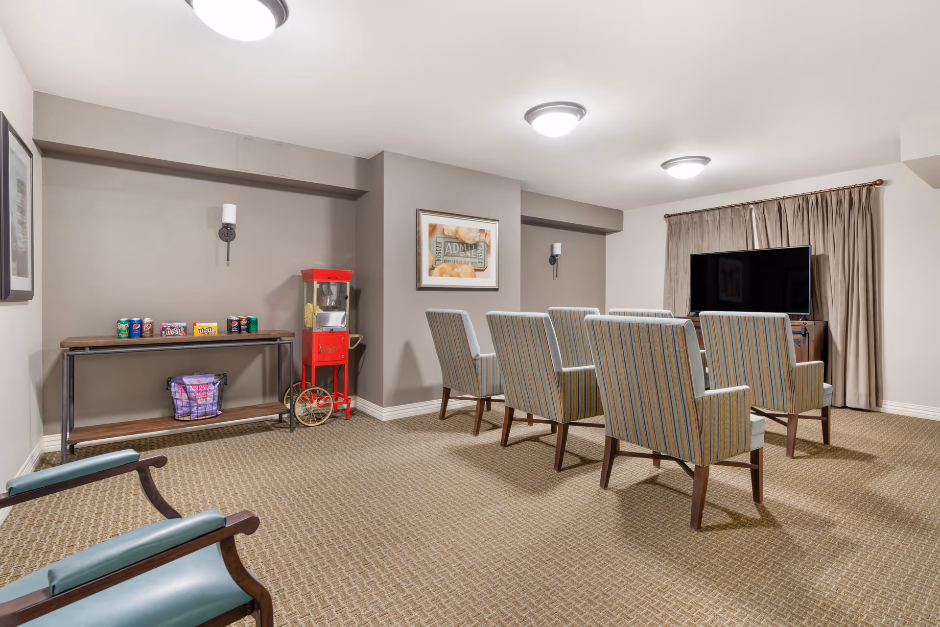 A small theater room with six striped upholstered chairs arranged in two rows facing a flat-screen TV. To the left, there is a wooden console table with assorted soda cans and candy boxes, and a vintage red popcorn machine next to it. The room has beige carpet, light gray walls, and ceiling lights, with a framed picture on the wall and beige curtains behind the TV.