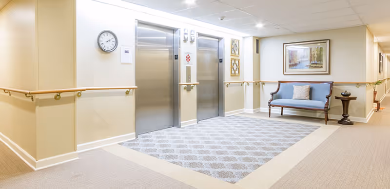 Interior hallway area with two stainless steel elevators, beige walls with wooden handrails, a blue cushioned bench with a pillow, a small round table with a decorative item, a wall clock, framed artwork, and patterned carpet in front of the elevators.