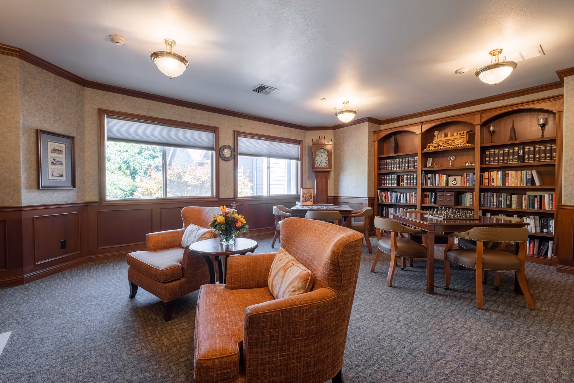 A cozy living room area in a senior living facility with two orange upholstered armchairs and a small round table with a flower arrangement. In the background, there is a wooden bookshelf filled with books and decorative items, a chessboard on a table with four chairs, and a grandfather clock near the windows. The room has large windows letting in natural light, beige walls with wooden wainscoting, and ceiling lights.