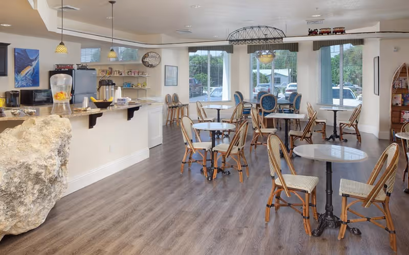 Bright communal dining area with multiple small round tables and wicker chairs, a serving counter at left, and large windows letting in daylight.