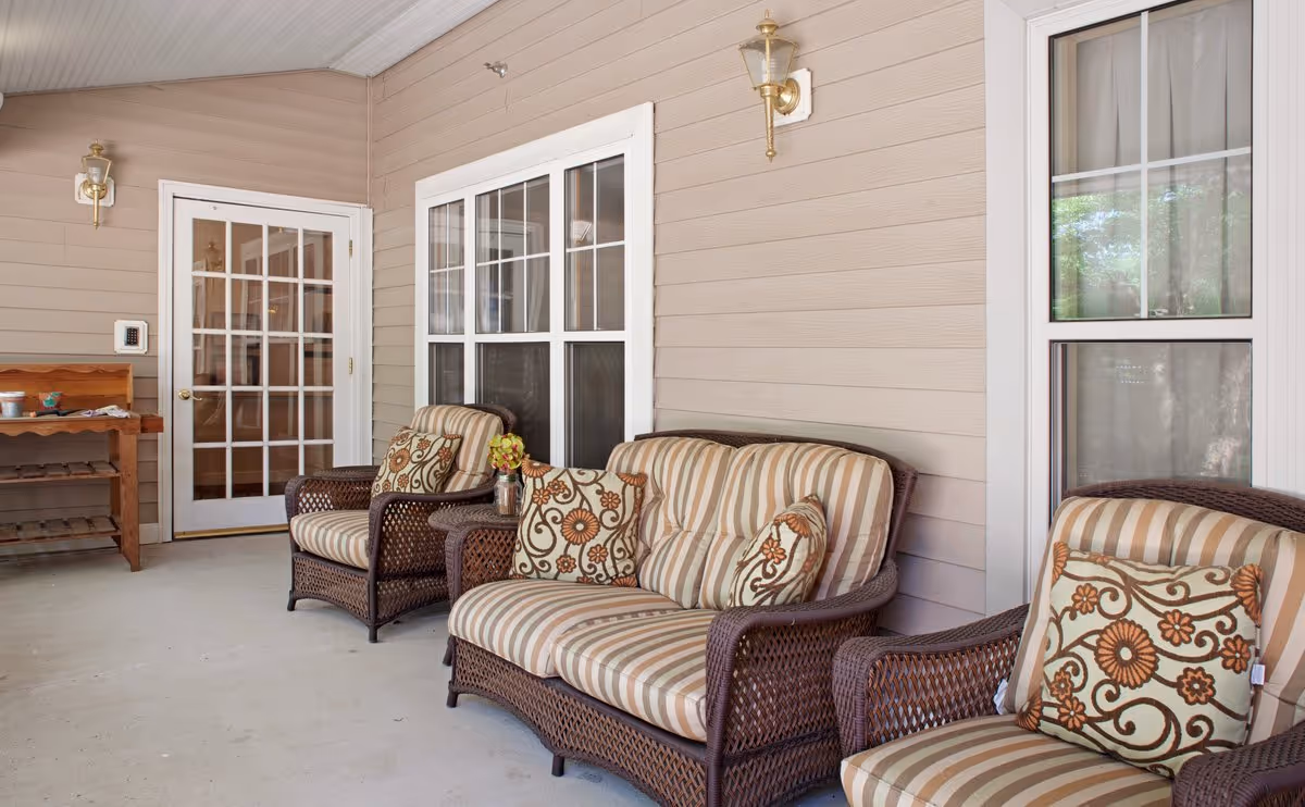 Covered porch with wicker seating and striped cushions arranged along a beige-sided wall.