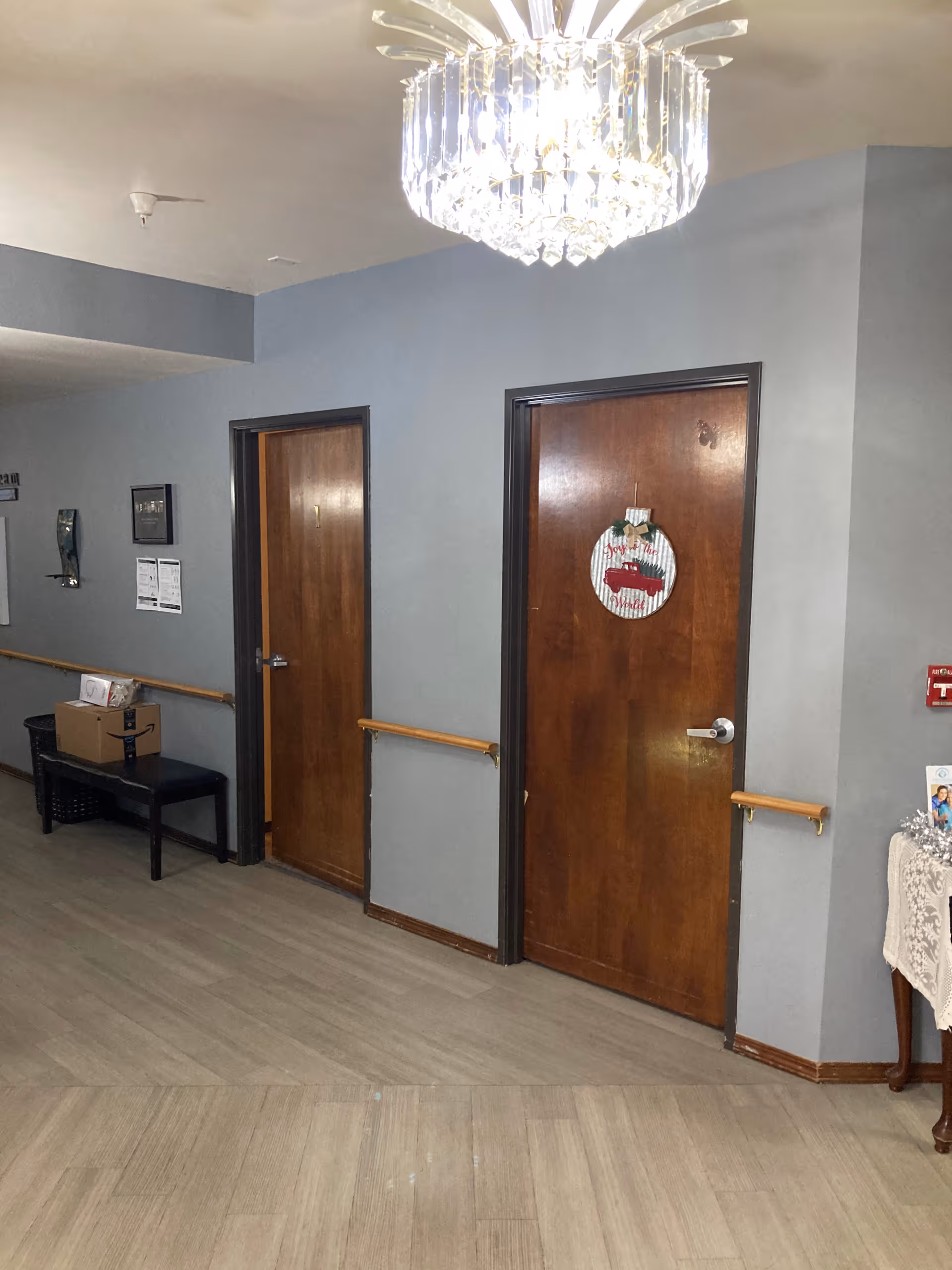 Hallway inside a senior living facility showing two wooden doors with handrails, a chandelier overhead, and a bench with boxes.