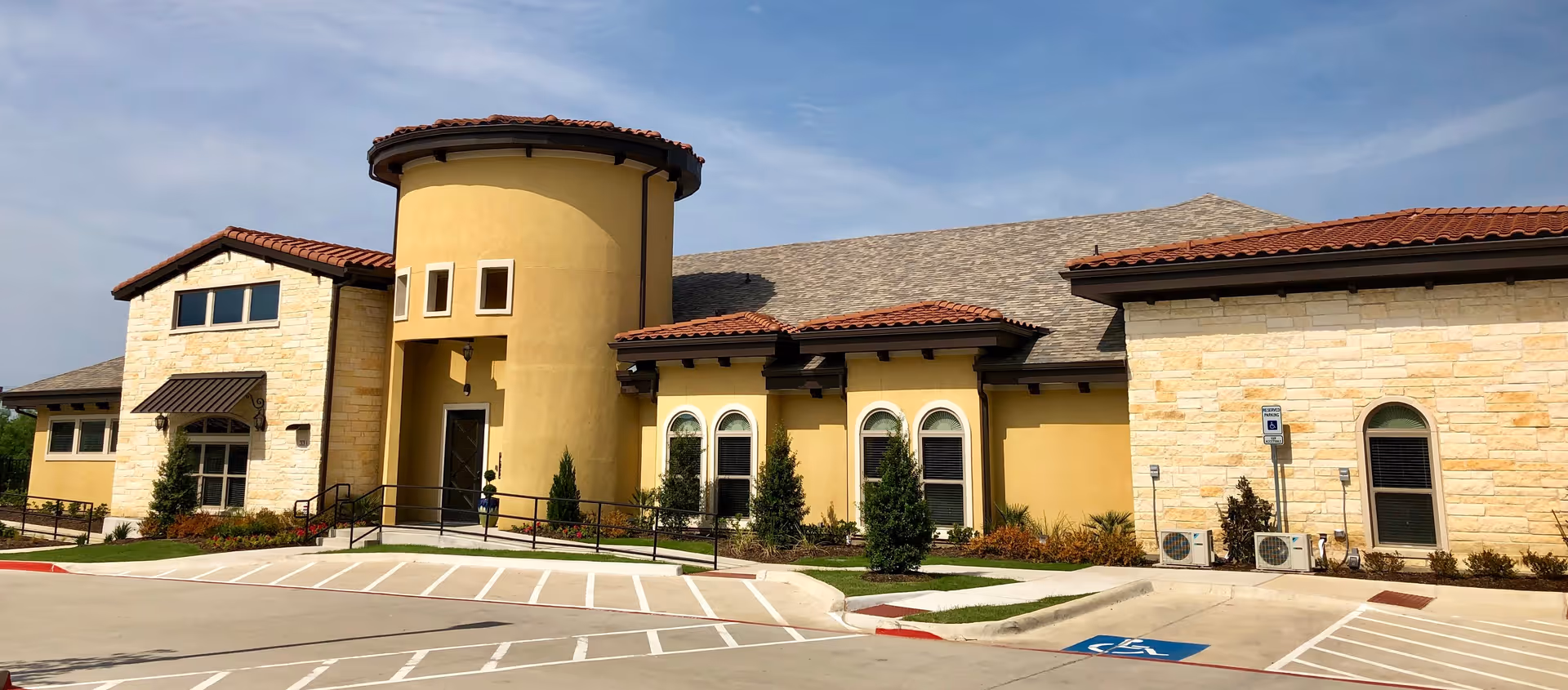 Exterior view of a senior living facility building with a yellow stucco and stone facade, red tile roof, arched windows, and a round tower-like entrance. The foreground shows a parking lot with marked parking spaces including a handicapped spot.