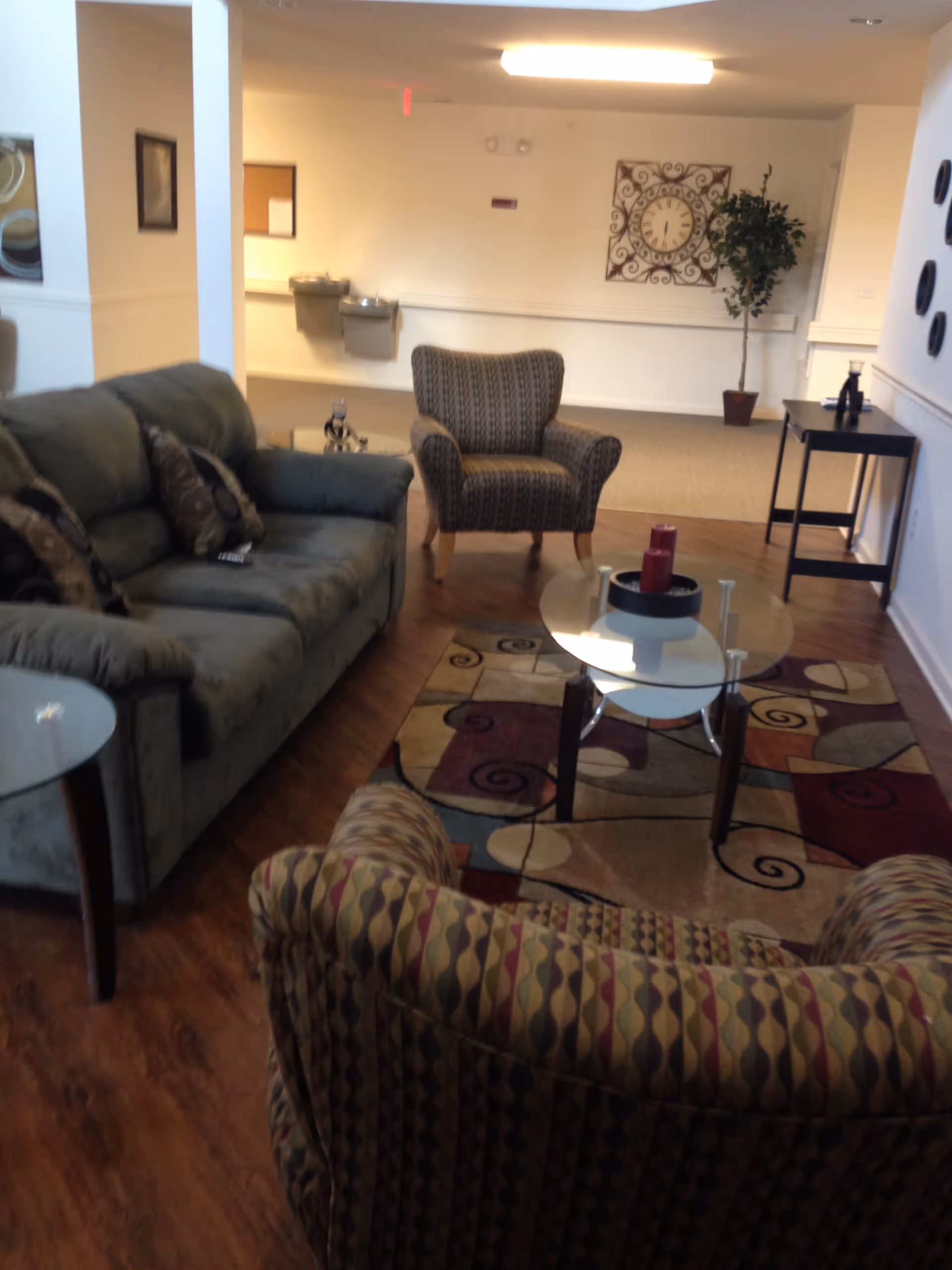 A furnished sitting area with a sofa, patterned armchairs, glass coffee table and decorative rug in a senior living facility.