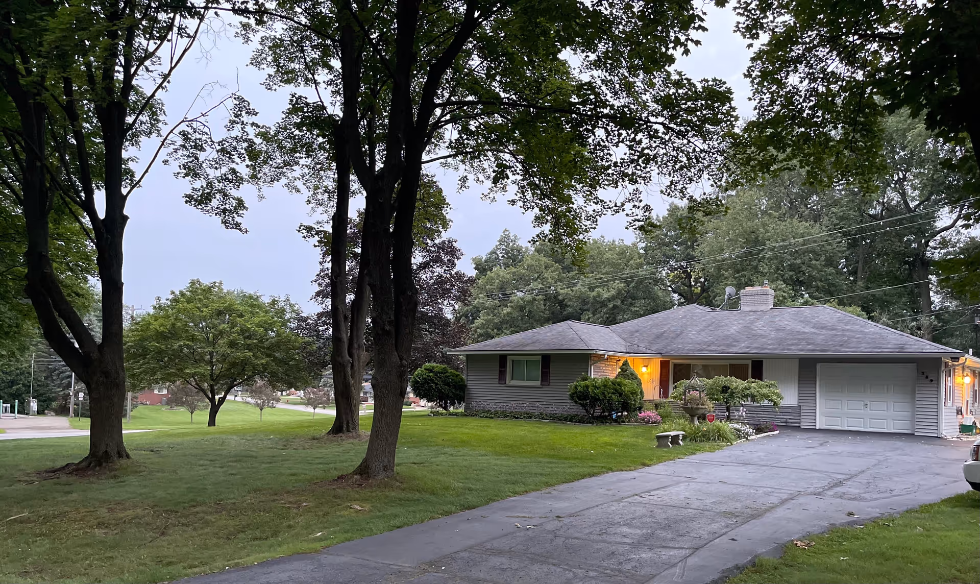 Single-story ranch-style house with a lit front porch, attached garage, driveway and a grassy yard with mature trees.