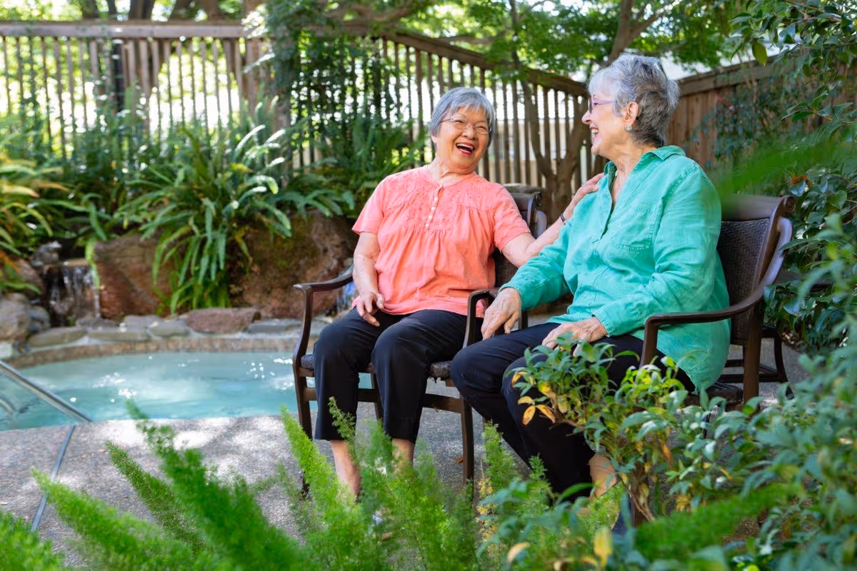 Two elderly women sitting on outdoor chairs by a small pool or hot tub, surrounded by lush greenery and a wooden fence, smiling and enjoying each other's company.