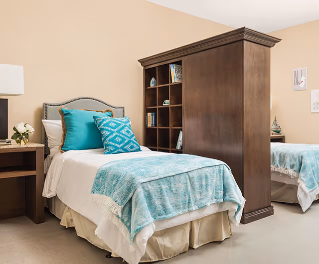 A bright shared bedroom with twin beds separated by a wooden shelving divider, each bed made with white linens and turquoise quilts and pillows.