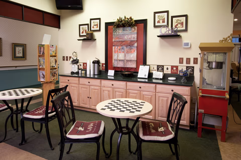A cozy cafe-style seating area in a senior living facility with two small round tables featuring checkerboard patterns, each surrounded by black metal chairs with maroon cushions labeled 'Cafe Espresso.' Behind the tables is a countertop with light wood cabinets, coffee dispensers, cups, and decorative items including framed pictures and a small statue. A red popcorn machine is visible to the right.
