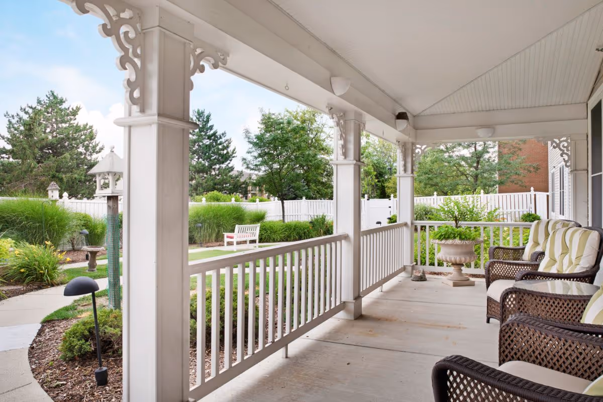 Covered porch area with white wooden railings and decorative trim, featuring cushioned wicker chairs and a large planter. The porch overlooks a landscaped garden with a white bench, birdhouse, and various green plants and trees under a partly cloudy sky.