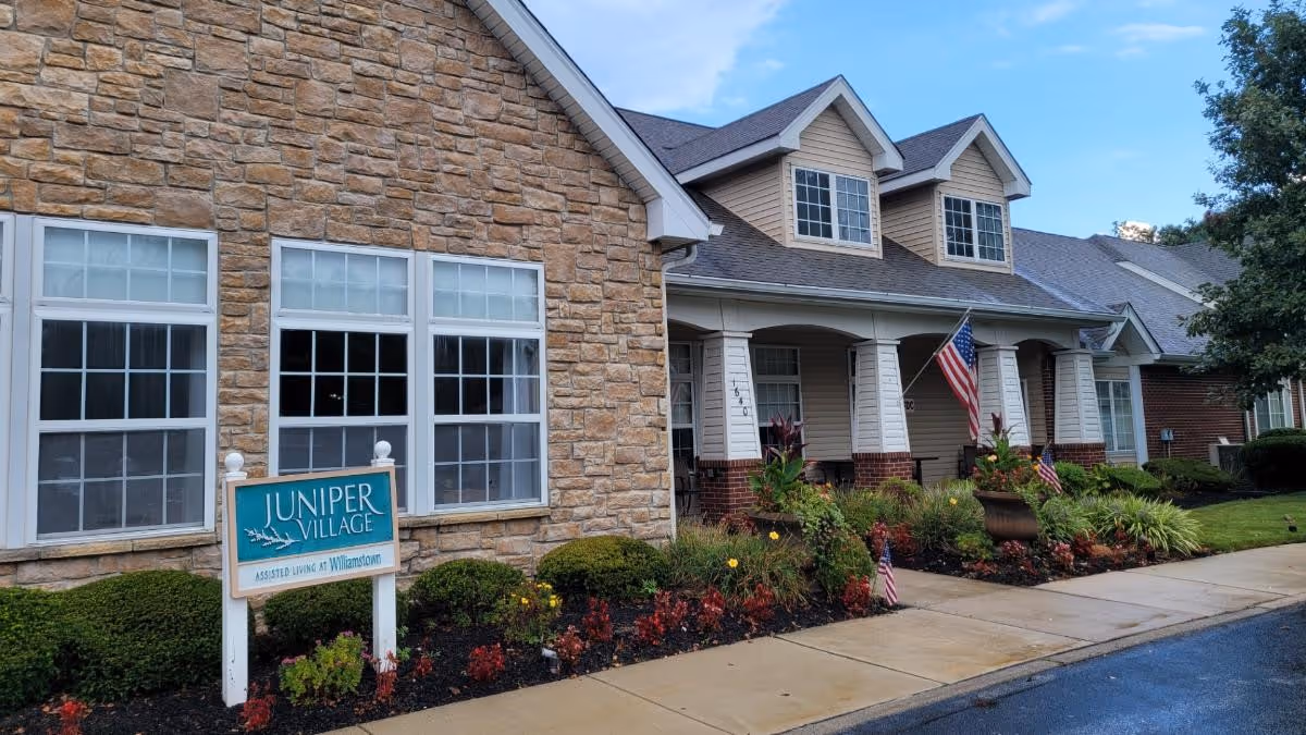 Front entrance of Juniper Village assisted living with stone and siding facade, a sign reading "Juniper Village", American flags, and landscaped walkway.
