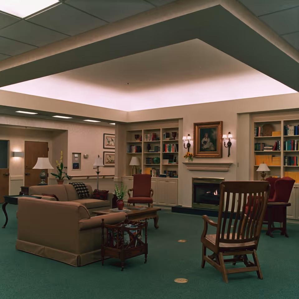 A cozy living room area with beige sofas, wooden chairs, a coffee table, and built-in bookshelves filled with books and decorative items. There is a fireplace with a framed painting above it and wall sconces on either side. The room has green carpet and soft overhead lighting.