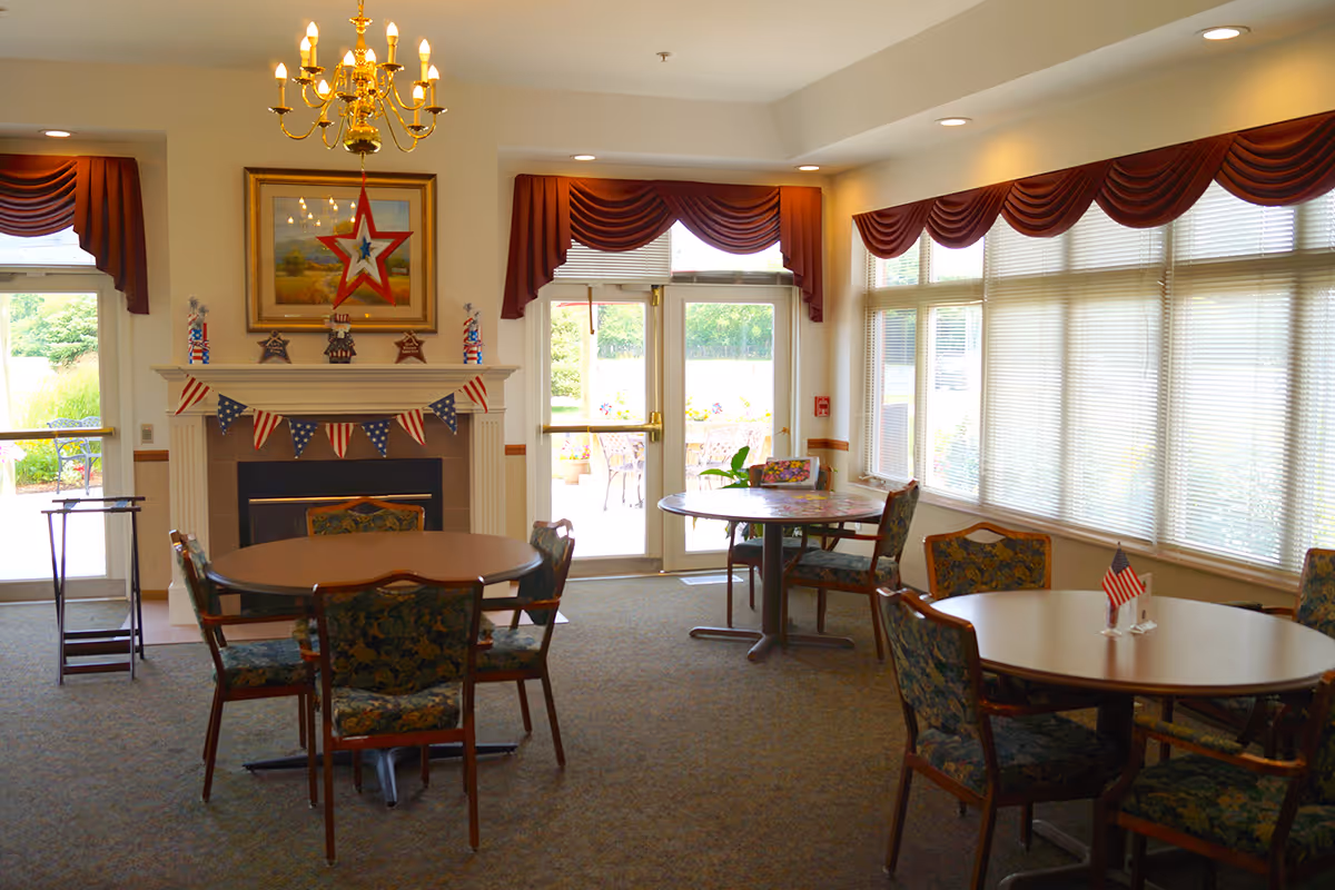 A bright common area in an assisted living center with round tables and floral upholstered chairs. The room features large windows with blinds and maroon valances, a fireplace decorated with patriotic bunting and star ornaments, and a gold chandelier hanging from the ceiling. Outside the glass doors, there is a patio with outdoor seating and greenery.
