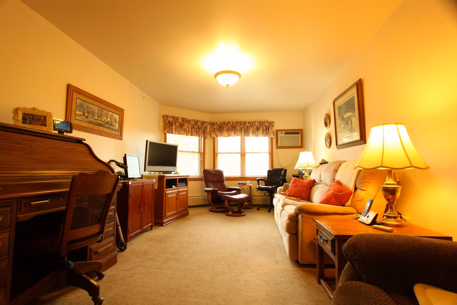 A cozy living room with beige walls and carpet, featuring a beige sofa with red cushions, a wooden side table with a lamp and a cordless phone, two armchairs near a window with floral valances, a TV on a wooden cabinet, and a wooden desk with a chair and framed pictures on the wall.