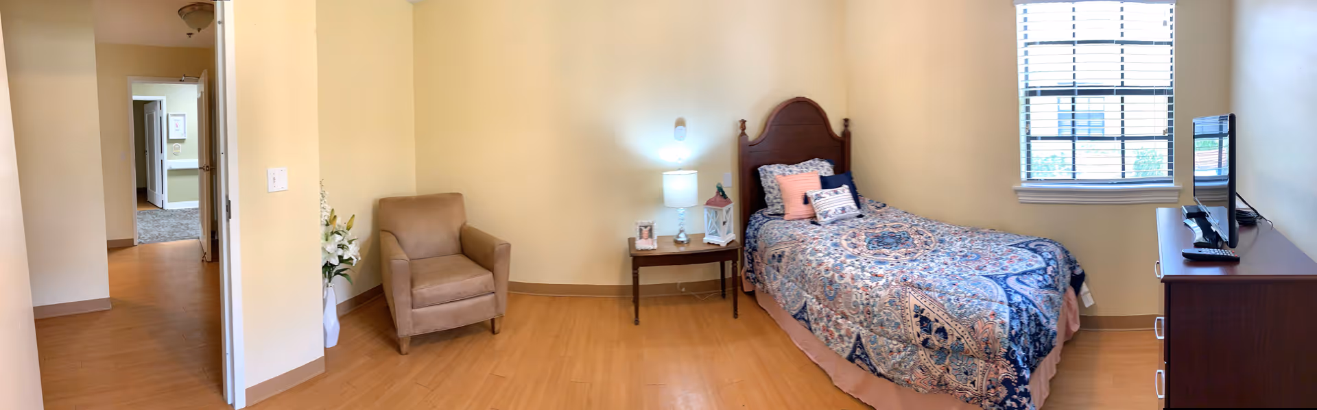 A cozy bedroom with a single bed covered in a patterned quilt, a wooden nightstand with a lamp and decorative items, a comfortable armchair, a window with blinds, and a wooden dresser with a television on top. The walls are painted light yellow and the floor has wood-like flooring.