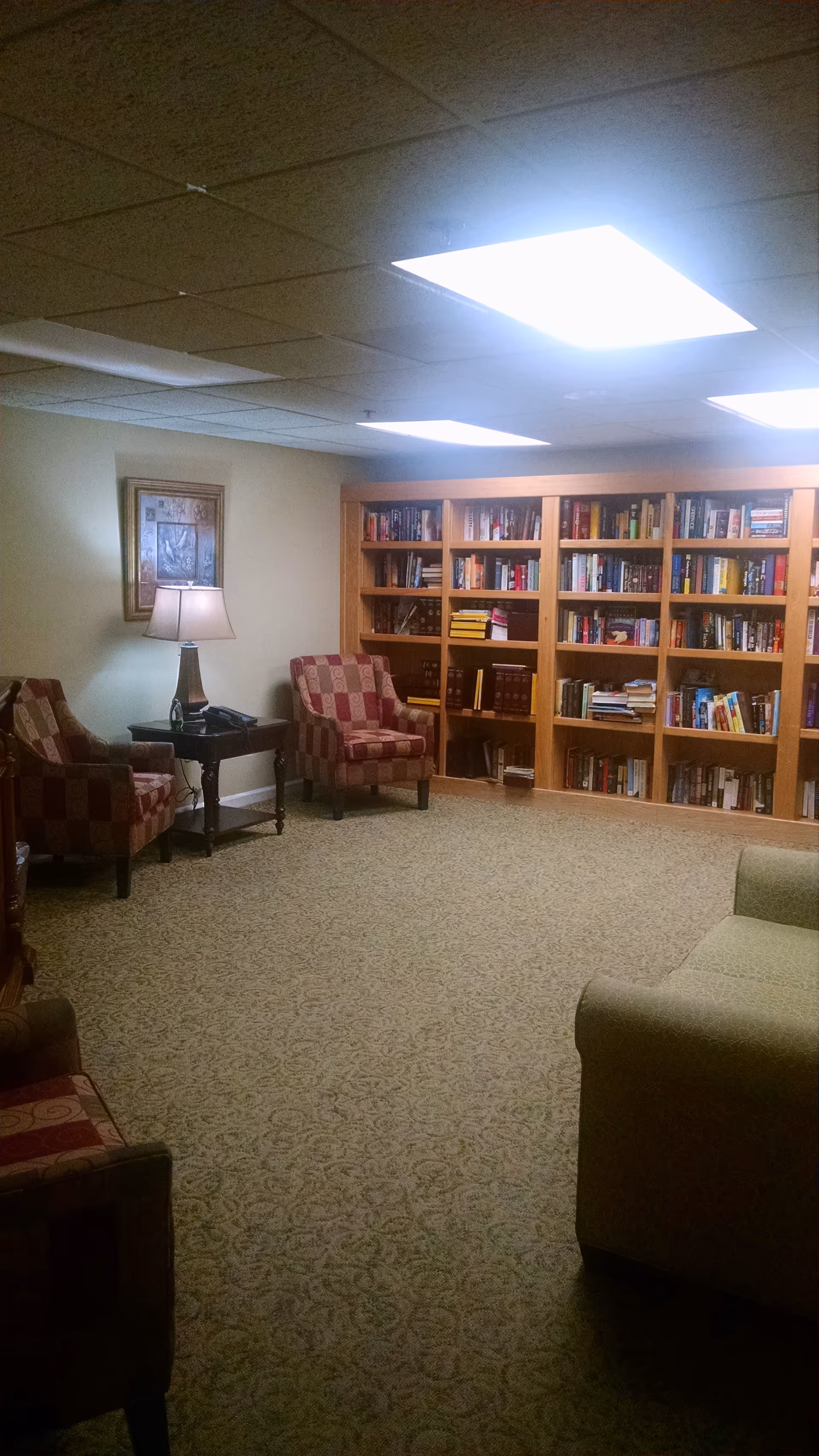 A cozy reading room with patterned armchairs, a green sofa, a wooden side table with a lamp and a phone, and a large wooden bookshelf filled with books. The room has a carpeted floor and a ceiling with bright fluorescent lights.