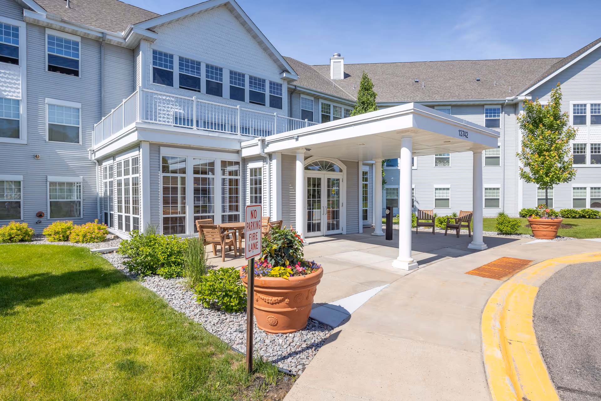 Front entrance of a white multi-story senior living building with a covered porte-cochere, potted plants and outdoor seating.