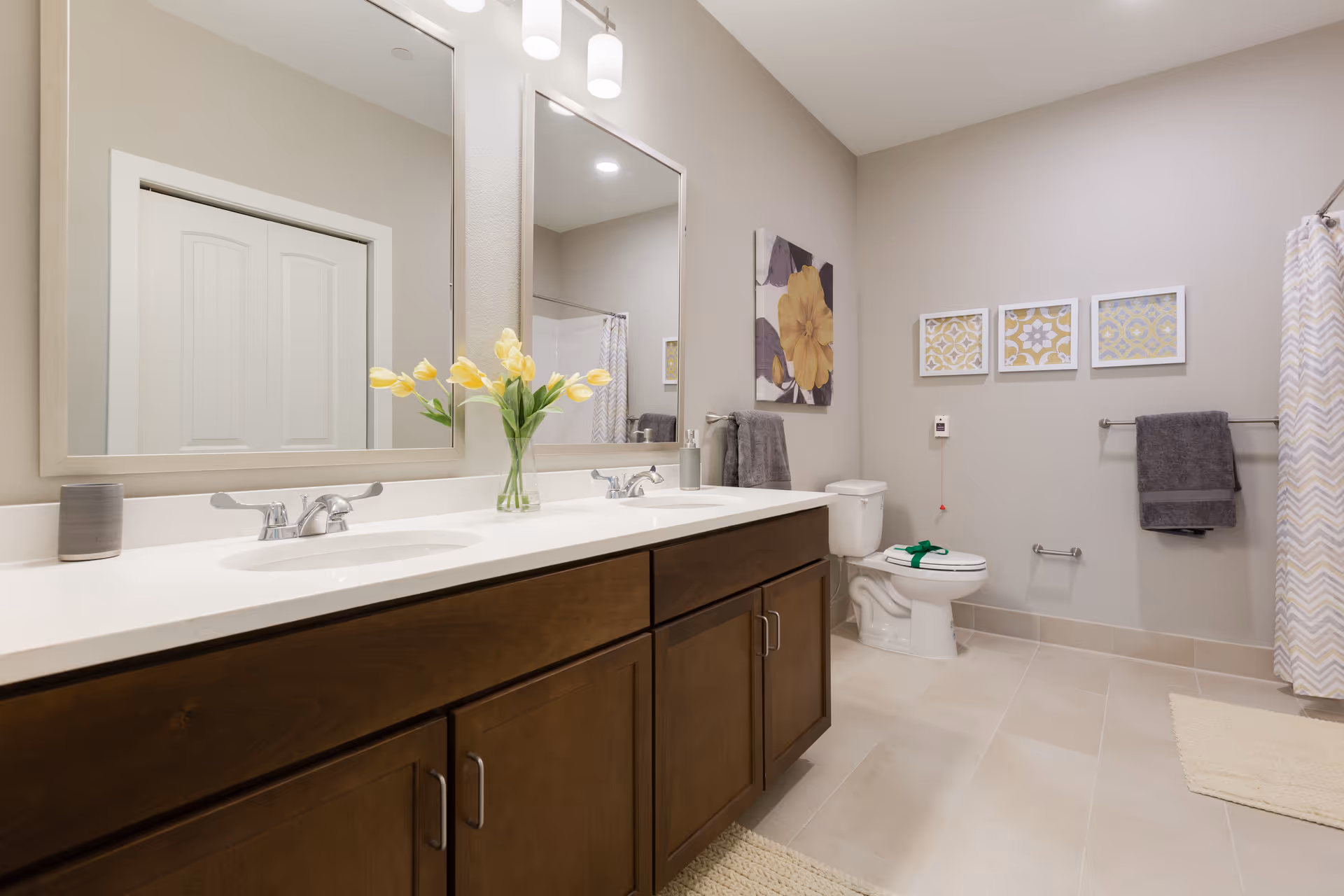 A spacious bathroom with a double sink vanity featuring dark wood cabinets and white countertops. Above the sinks are two large mirrors with three light fixtures. A vase with yellow tulips is placed on the countertop. The bathroom has beige tiled floors, a toilet with a green ribbon on the lid, a towel rack with a dark gray towel, and a shower with a chevron-patterned curtain. The walls are decorated with three framed artworks and a large floral painting.