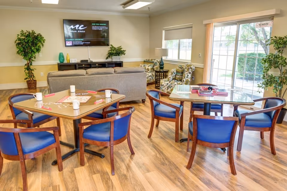 A bright and spacious common area in a senior living facility with two tables and blue cushioned chairs arranged around them. One table has a Scrabble game set up with tiles and mugs. In the background, there is a gray sofa facing a wall-mounted TV, two floral armchairs with a small side table and lamp between them, and large windows with blinds and a sliding glass door letting in natural light. Potted plants are placed around the room, and the floor is wood-patterned.