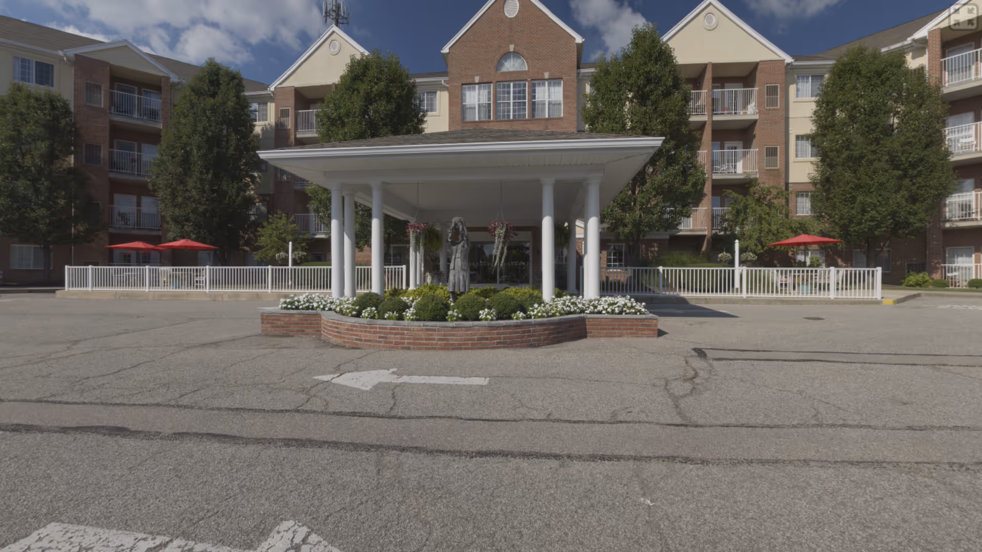 Front exterior view of Barrington of Fort Thomas facility showing a covered entrance with white columns, a brick planter with flowers and a statue, surrounded by a multi-story building with balconies and trees.