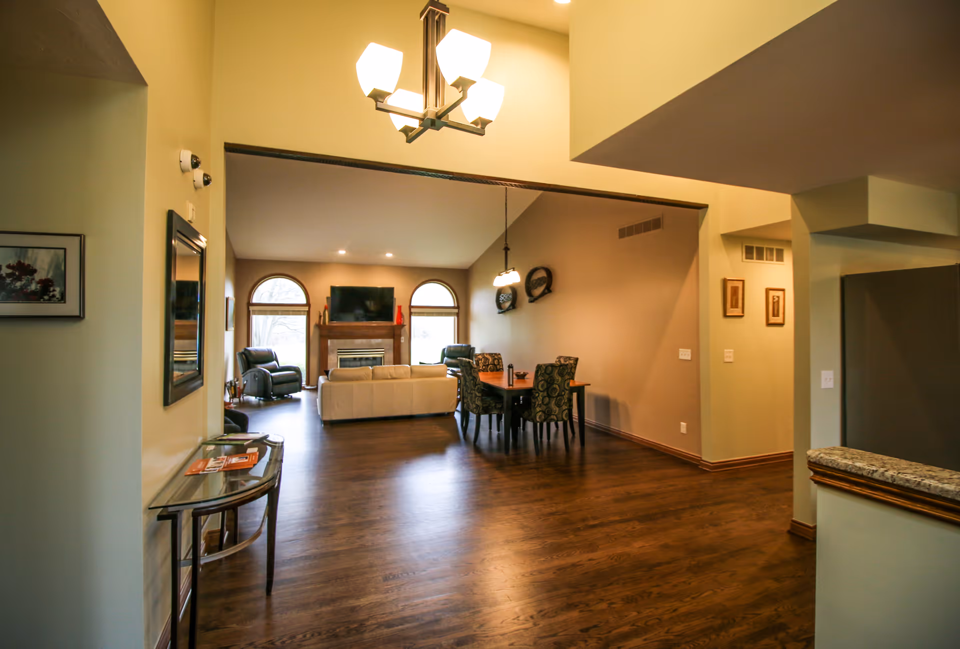 Interior view of a spacious living and dining area with wooden flooring. The living room features a white sofa, two recliners, a fireplace with a TV mounted above it, and two arched windows. The dining area has a wooden table with four patterned chairs and a hanging light fixture above it. The walls are painted in neutral tones, and there is a small glass table with magazines near the entrance.