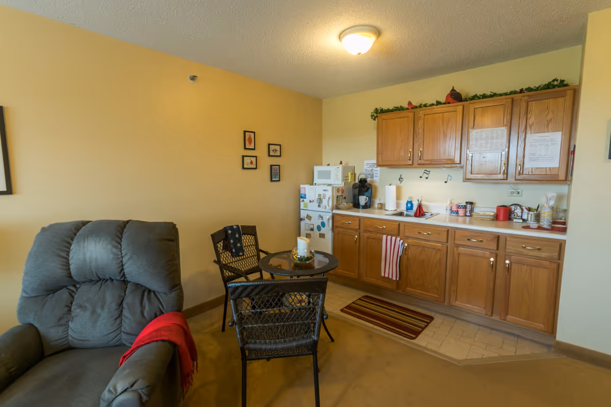 A cozy kitchenette area in a senior living facility with wooden cabinets, a small white refrigerator, microwave, coffee maker, and a sink. There is a small round glass table with two wicker chairs nearby. A comfortable gray recliner chair with a red throw blanket is positioned to the left. The walls are painted light yellow and decorated with small framed pictures and musical note decals above the sink. The floor has a combination of carpet and tile with a striped rug in front of the sink.
