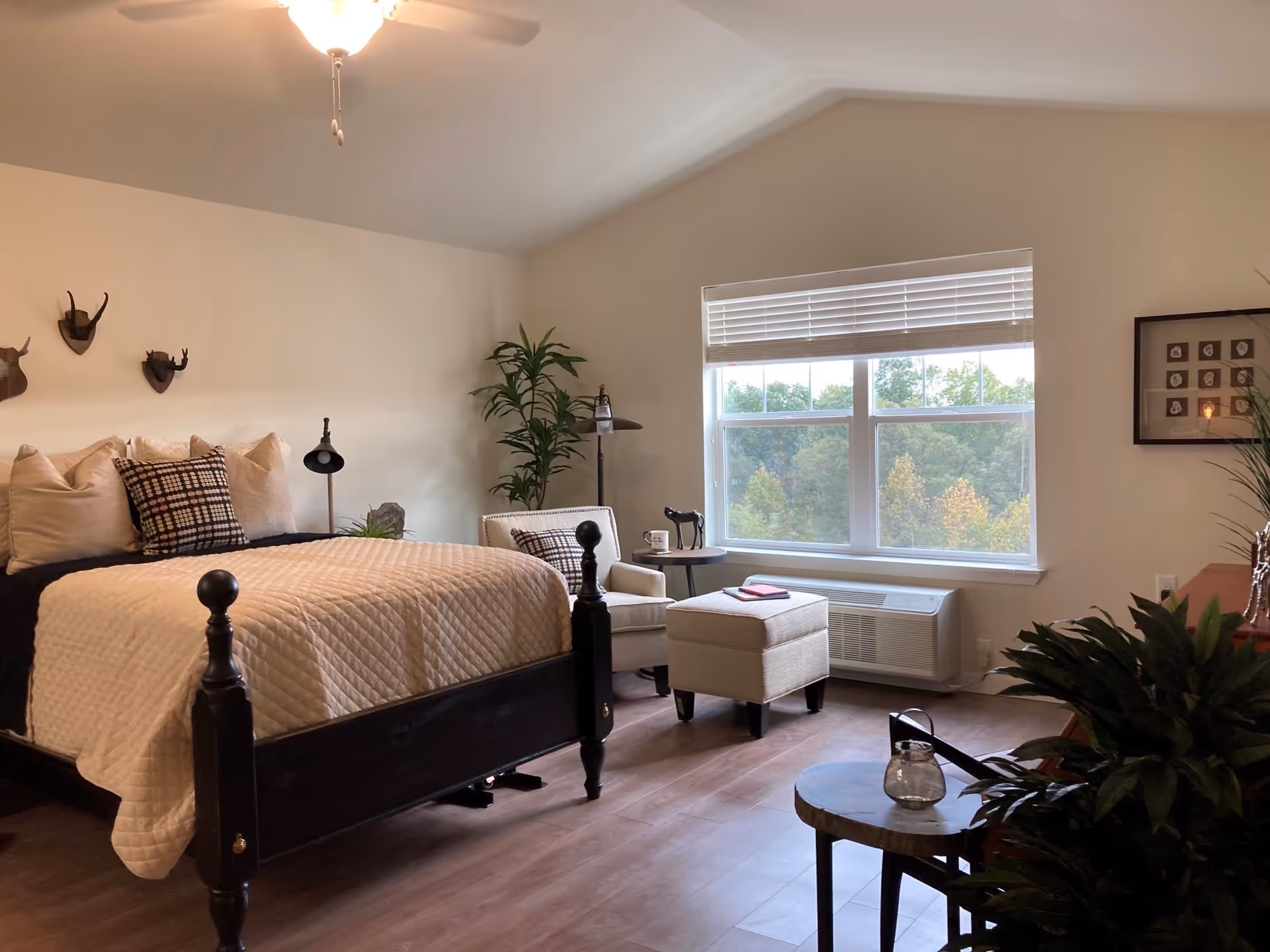 A cozy bedroom with a black wooden bed frame, beige quilted bedding, and multiple pillows. There is a beige armchair with a matching ottoman near a large window with white blinds, showing a view of trees outside. The room has wooden flooring, a ceiling fan with a light, several decorative plants, and wall decorations including mounted antlers and framed pictures.